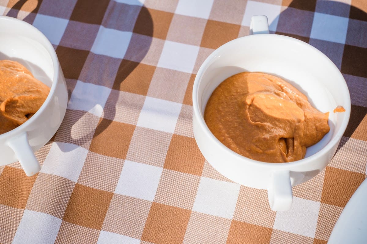 Two white cups of orange-colored romesco sauce on a checkered tablecloth.