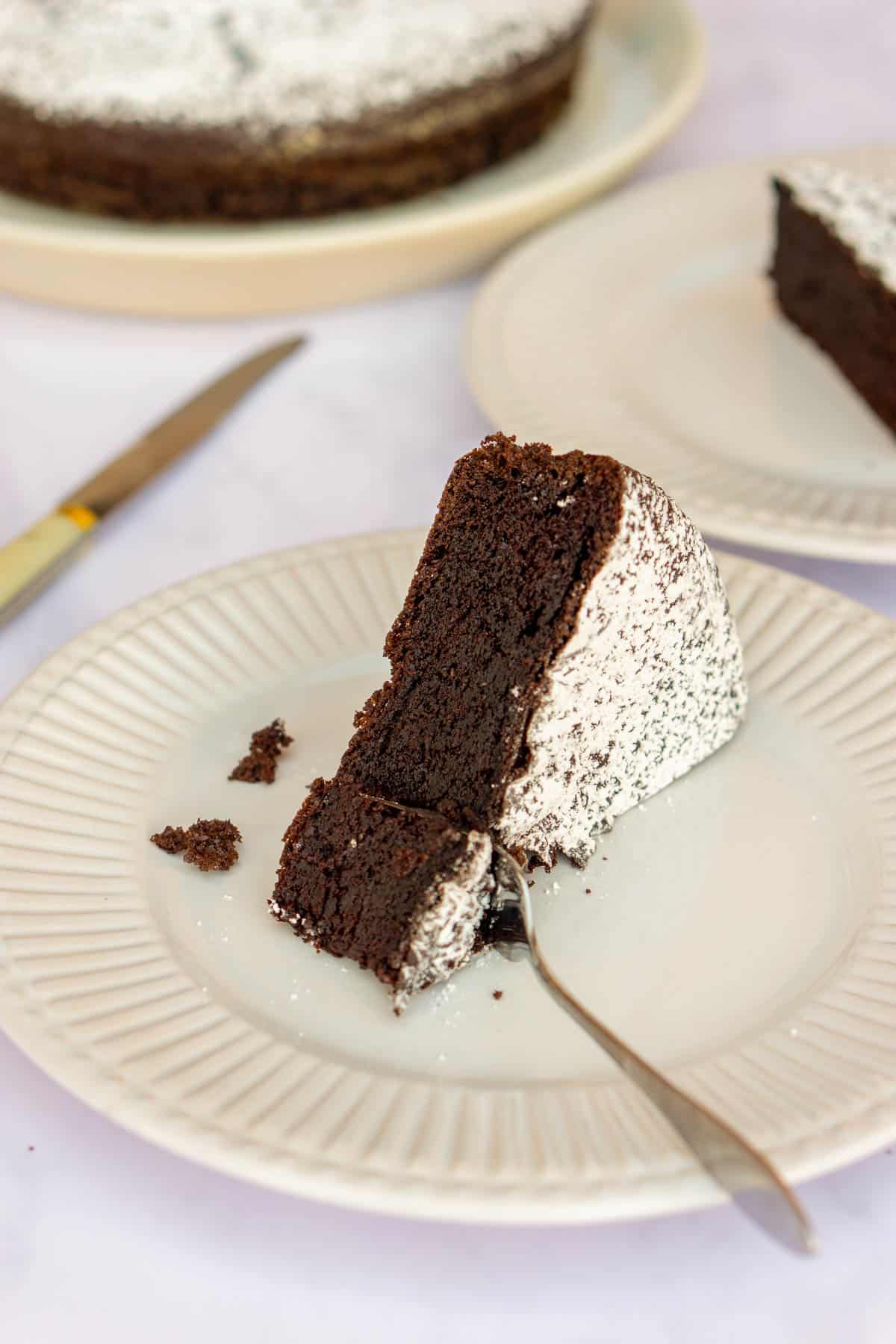 Slice of chocolate olive oil cake on a white plate with more cake in the background