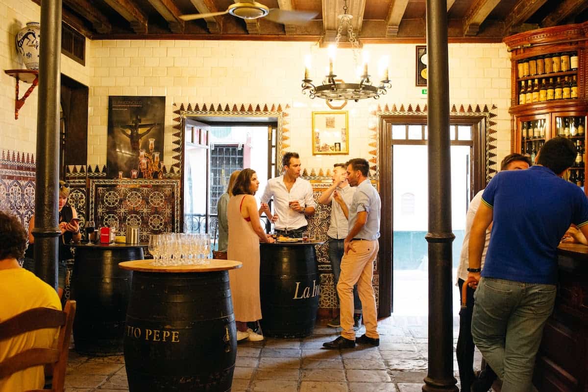 Interior of a bar with tiled walls and people standing around barrels to eat and drink.