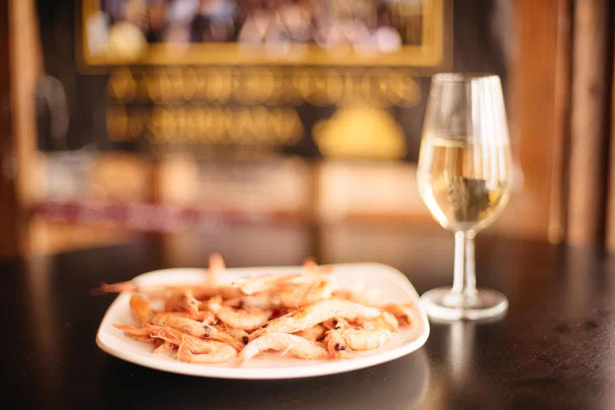 Boiled shrimp on a plate next to a glass of pale yellow sherry wine.