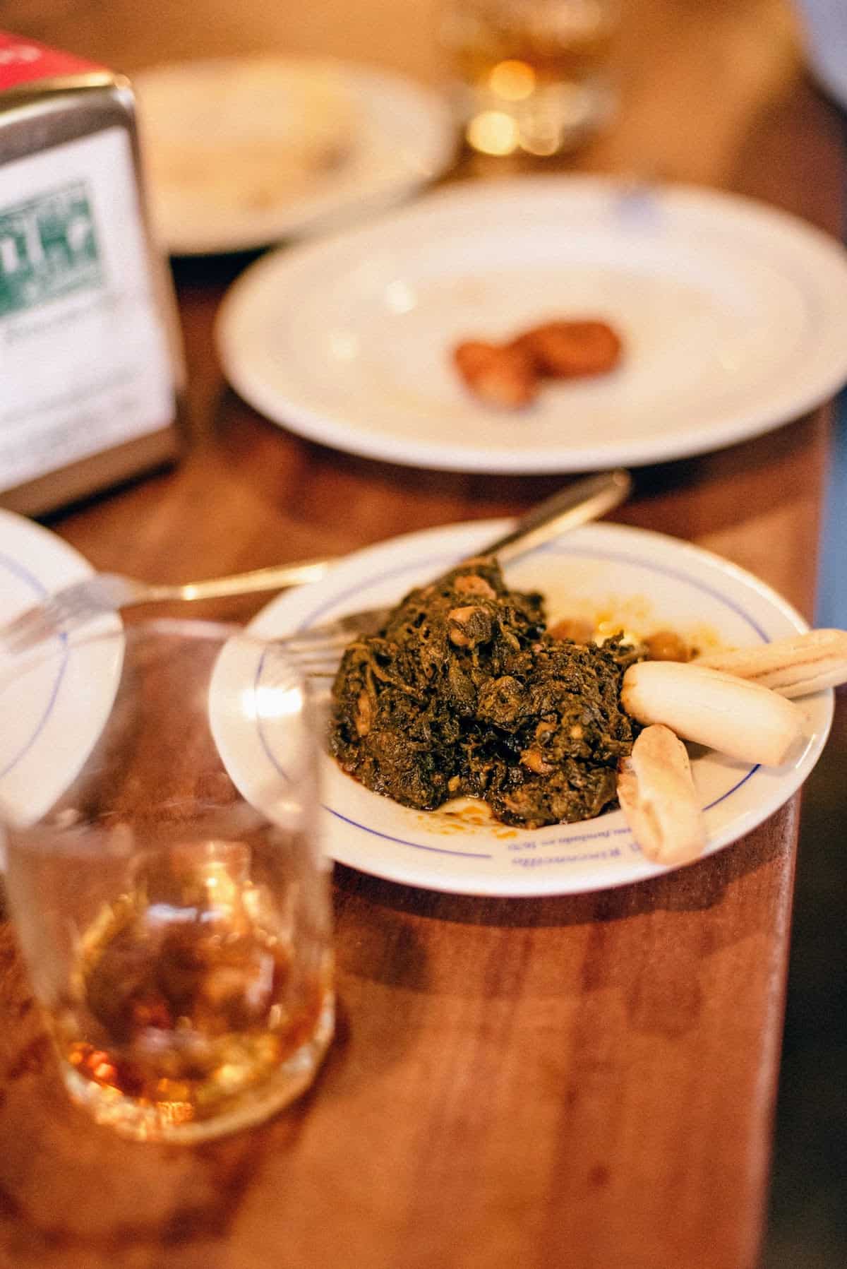 Plate of spinach and chickpea stew next to a glass of vermouth on a wooden tabletop.