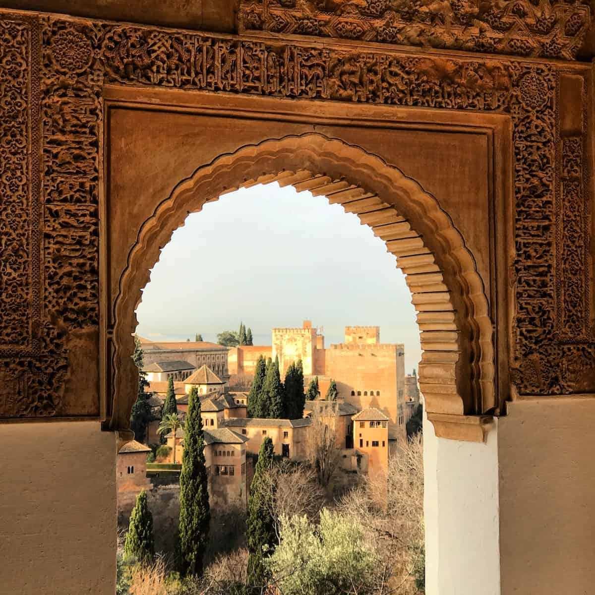 Complex of large brown buildings seen through a stone arch decorated with Moorish details.