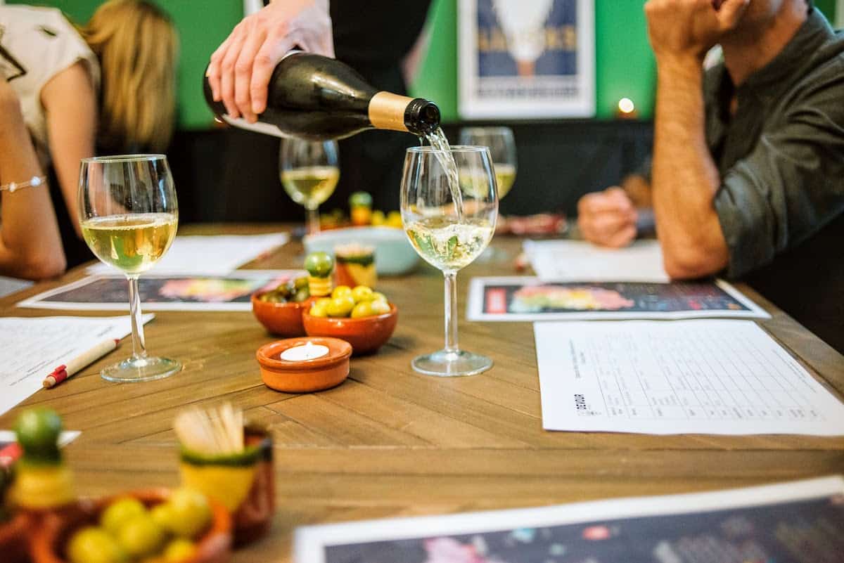 Close up of a person pouring a glass of white wine, with several other glasses of wine and dishes of olives visible on the table.