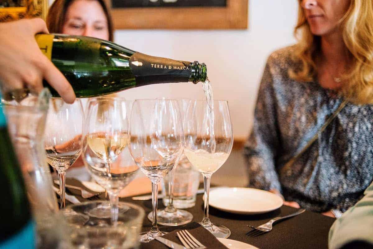 Several glasses of sparkling wine being poured with two women seated in the background.