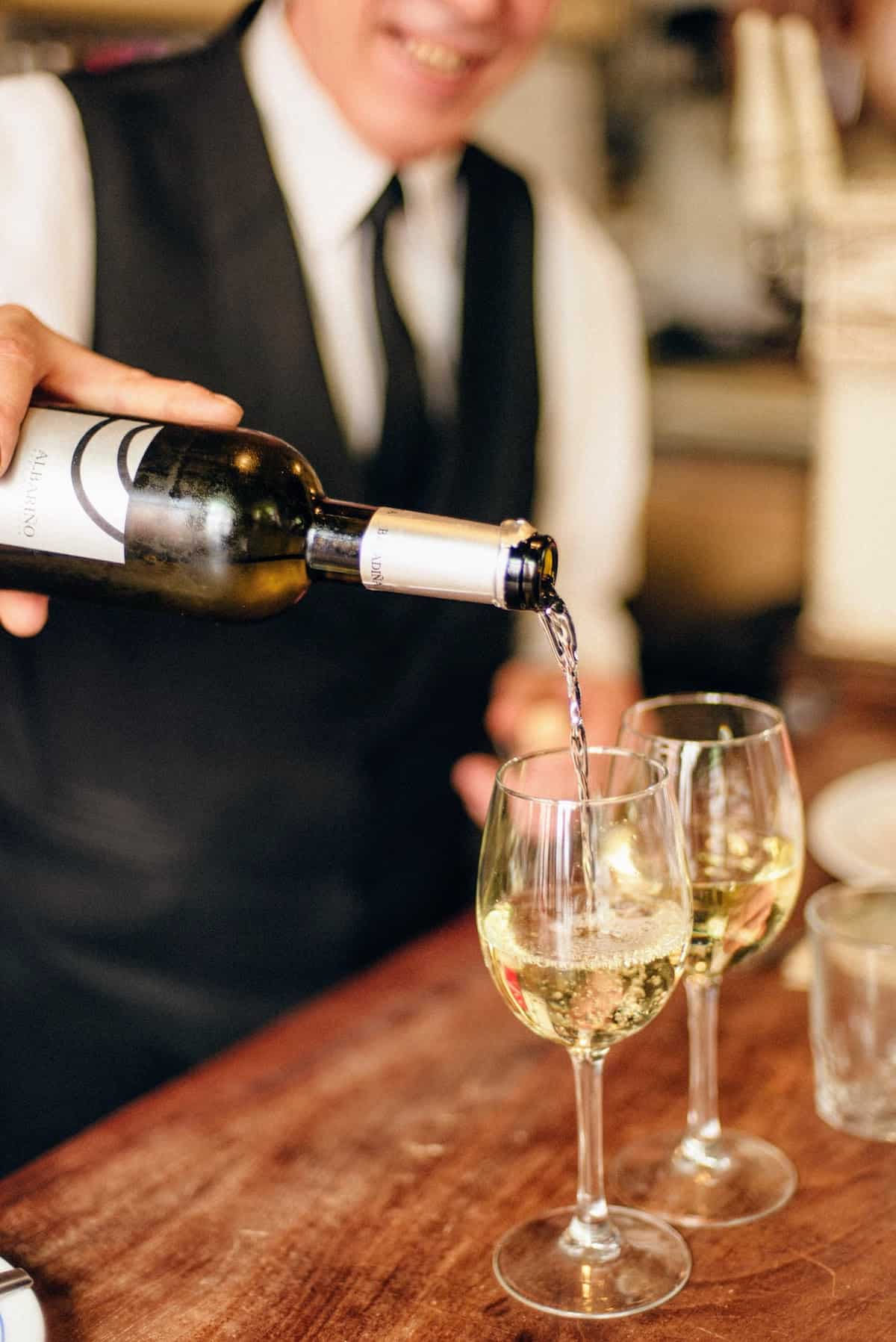 Close up of a bartender pouring two glasses of white wine on a wooden bar top.