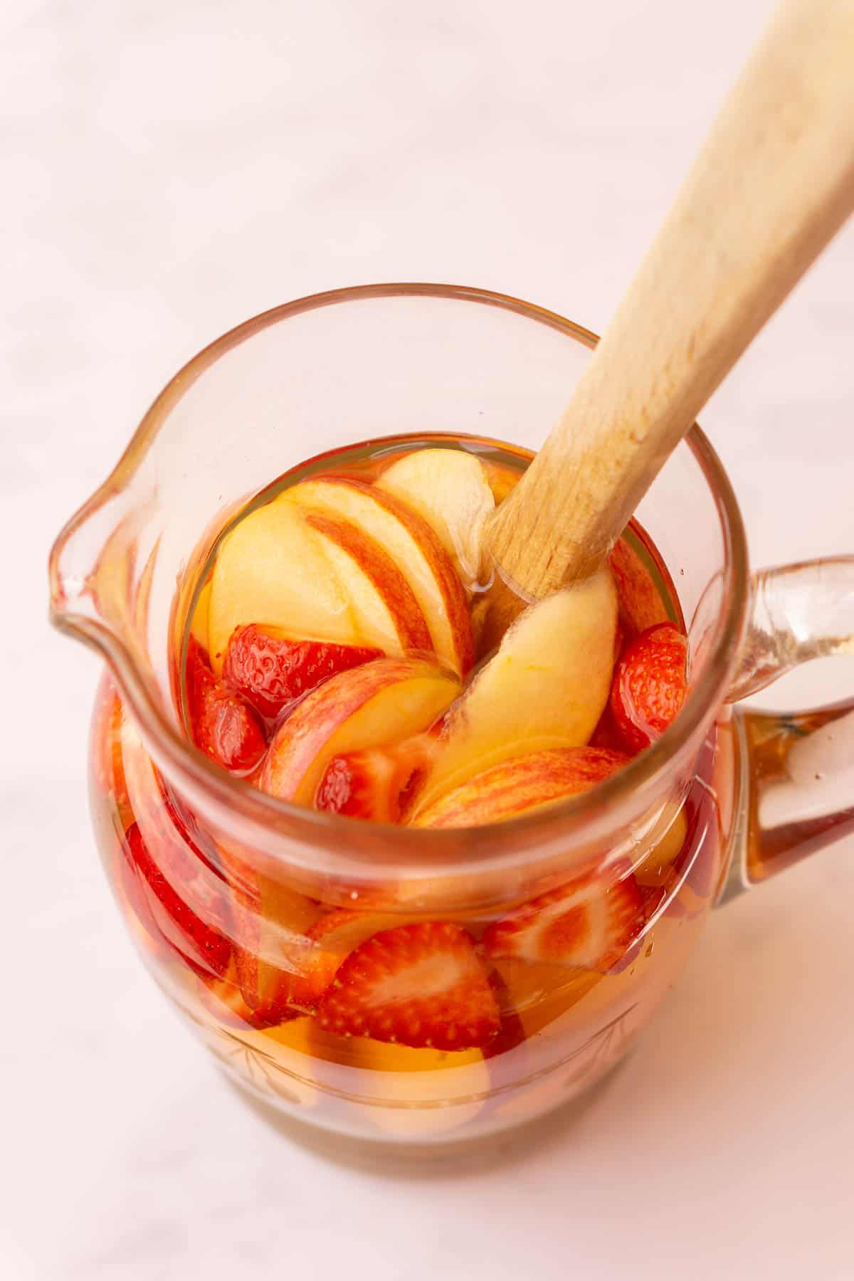 Pitcher of white wine sangria filled with different fruits and with a wooden spoon.
