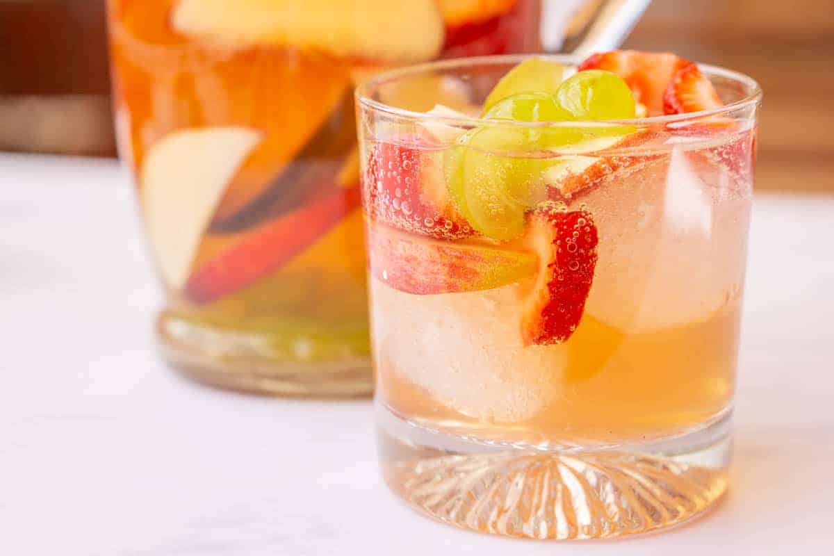 Delicious white wine cocktail with fruit in a glass with ice cubes and the pitcher behind it.