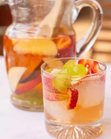 Delicious white wine sangria in a glass with ice cubes and the pitcher behind it.