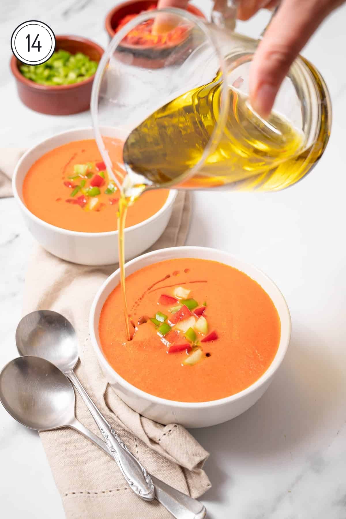 Step 14 gazpacho recipe - a bowl of gazpacho with chopped veggies and a hand pouring olive oil. 