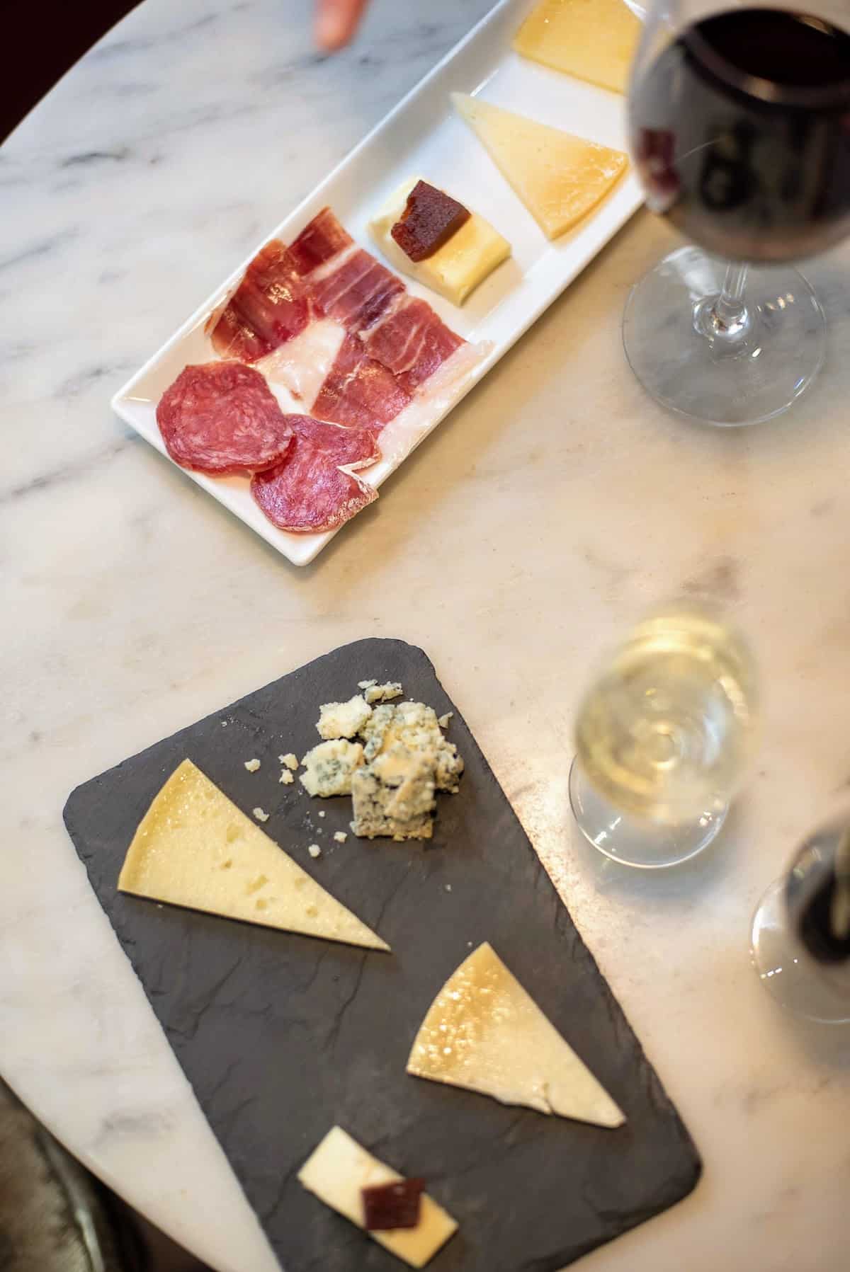 Overhead shot of small cheese and charcuterie trays beside two wine glasses on a marble tabletop.