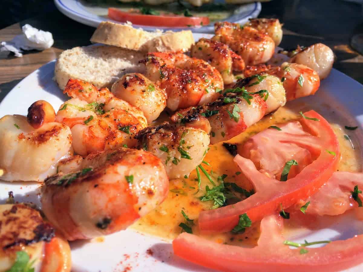 Shrimp skewers coated in spices and herbs on a white plate with sliced tomatoes and bread.