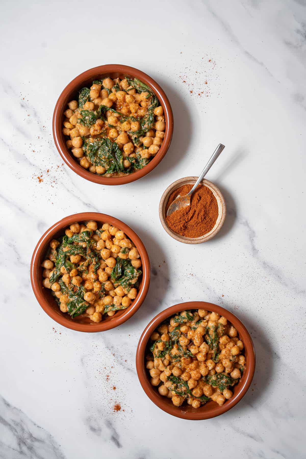 Overhead view of three small clay bowls of spinach and chickpeas with a dish of paprika.