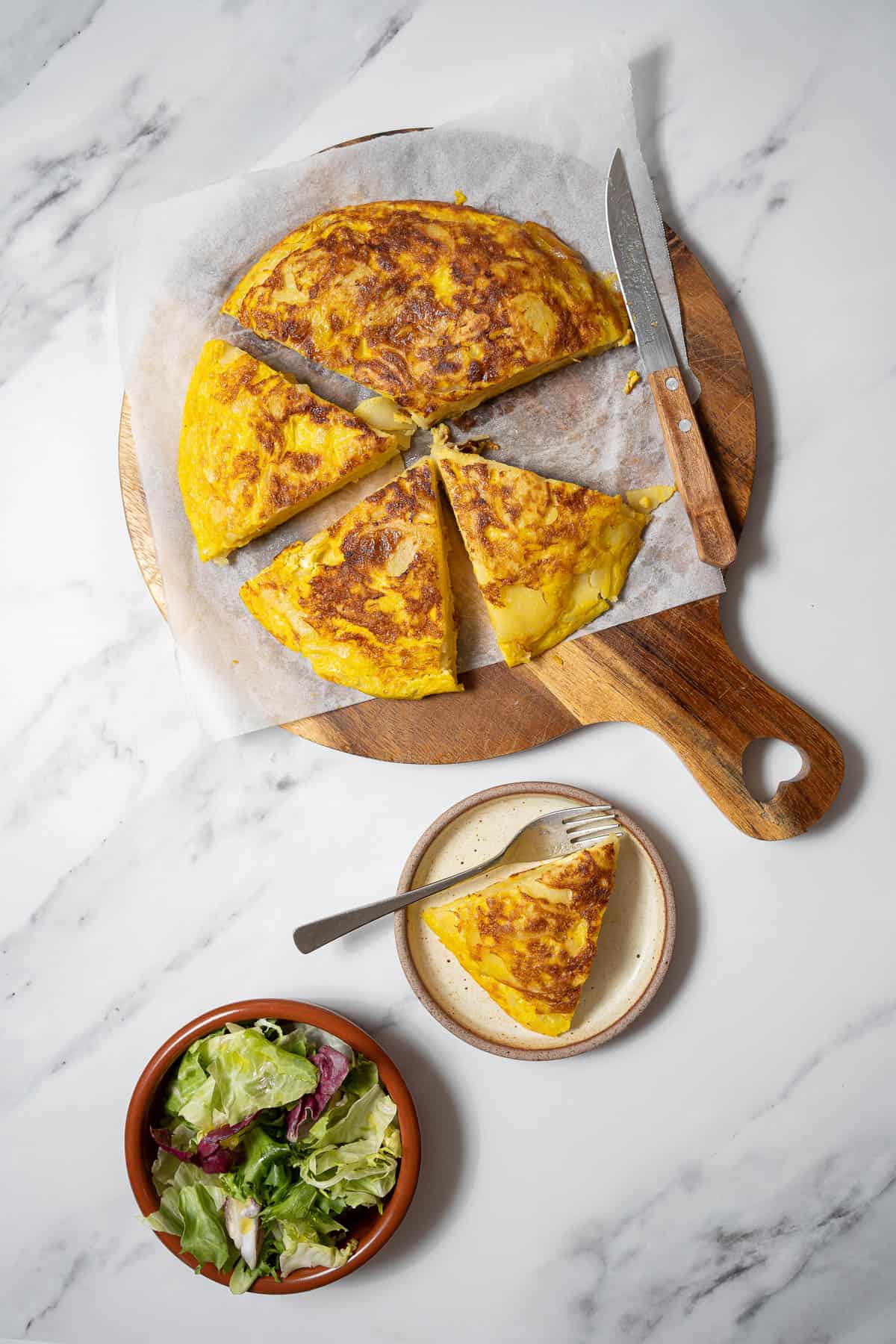 tortilla de patata on a cutting board with a bowl of lettuce.