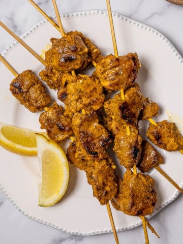 several skewers of pinchos morunos on a plate with lemon wedges.