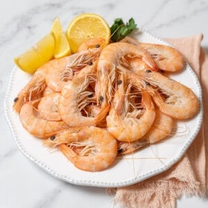 boiled shrimp on a white plate with lemon slices and a sprig of parsley.