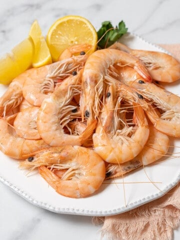 boiled shrimp on a white plate with lemon slices and a sprig of parsley.