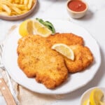 plate of chicken milanesa with lemon wedges and french fries.